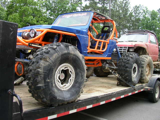 Blue off-road vehicle with orange roll cage and large tires on a trailer, next to a red pickup truck
