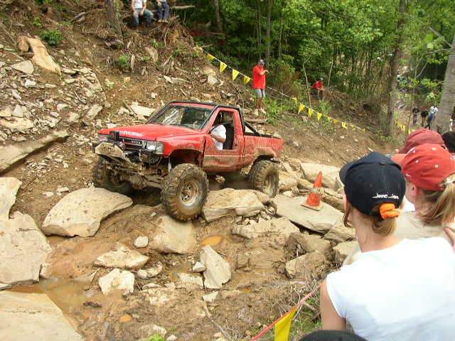 Red Toyota pickup truck with large tires climbing over rocks on a muddy trail