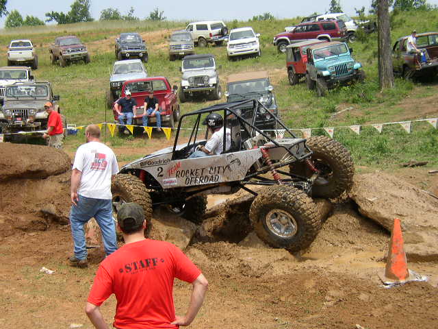 Off-road buggy with 'ROCKET CITY OFFROAD' on its side, climbing a dirt mound. Several other off-road vehicles are parked on a grassy hill in the background