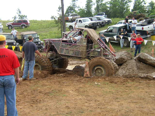 Custom off-road buggy stuck in mud pit with people watching and other vehicles in background