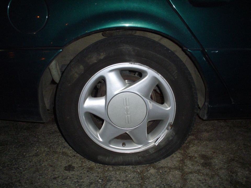 Close-up of a dark green car's rear wheel and fender. The wheel has a silver five-spoke hubcap with a logo