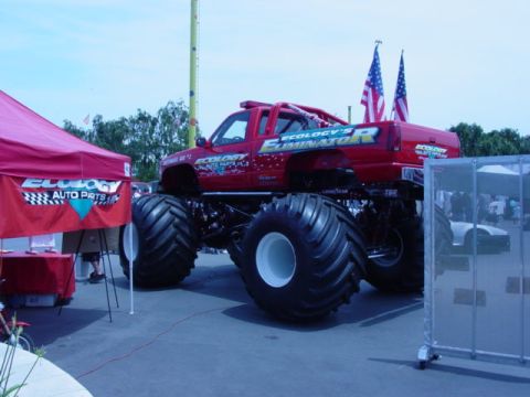 1992 Ford Bronco - A large red monster truck with custom graphics is displayed outdoors next to a promotional tent.