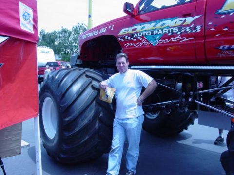 1992 Ford Bronco - A man stands next to a red monster truck with "Ecology Auto Parts" branding and oversized tires at an off-road show.