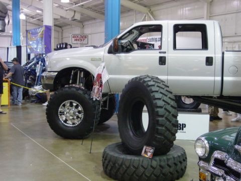 1992 Ford Bronco - A heavily modified pickup truck with oversized tires is showcased at an event, with other vehicles and displays visible in the background.