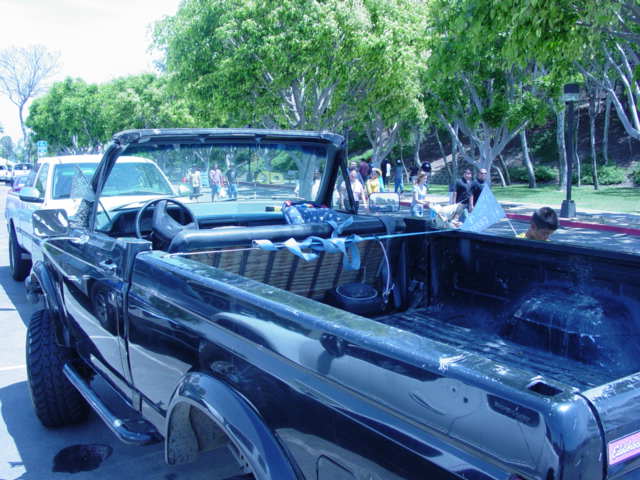 Black Ford Bronco with top removed, viewed from rear quarter, parked near a park with trees and people