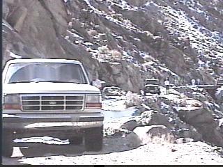 1993 Ford Bronco - A white SUV is parked on a rocky, snow-dusted mountain road, with another vehicle visible further up the road.