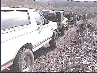 1993 Ford Bronco - Several off-road vehicles are parked in a line on a dirt road with people standing nearby.