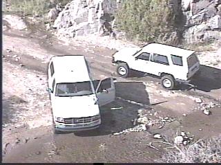 1993 Ford Bronco - Two white SUVs navigate a dirt road in a rocky, outdoor setting. Super Motors logo visible.