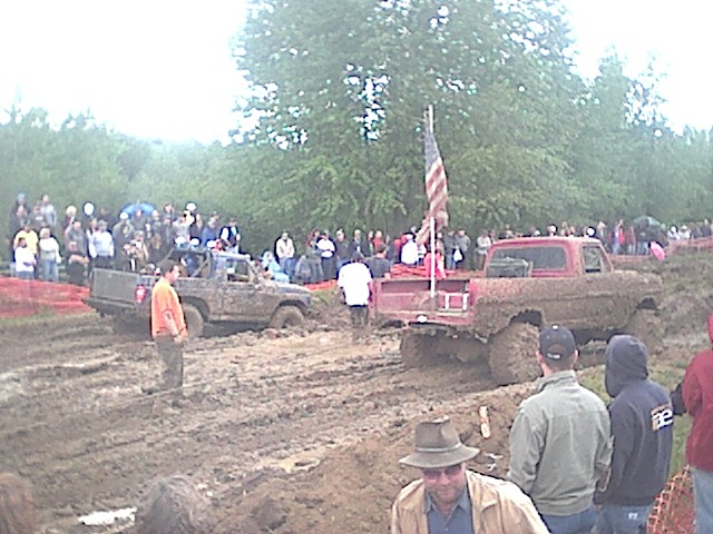 Red lifted pickup and dark truck in a muddy mud-bog pit with crowd and US flag