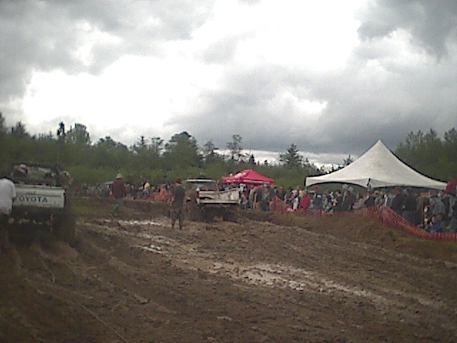 Trucks competing in a muddy off-road event with spectators and event tents