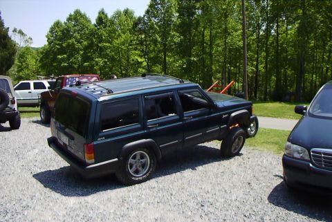 1995 Ford Bronco - A dark green Jeep Cherokee XJ is parked on a gravel lot with other vehicles and trees in the background.