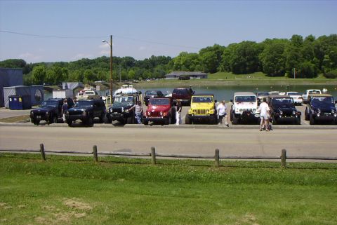 1995 Ford Bronco - A collection of Jeeps are gathered in a parking lot adjacent to a lake on a sunny day.