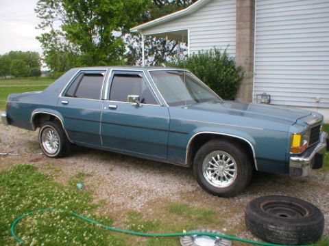 1987 Ford Crown Victoria LTD - This blue sedan is parked in front of a house with a spare tire nearby.