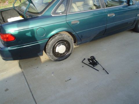 1993 Ford Taurus - A green sedan is parked on a concrete surface with its trunk open and a flat tire, accompanied by a jack and lug wrench.