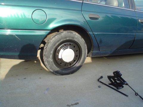 1993 Ford Taurus - A green 1993 Ford Taurus sedan is parked on concrete with a flat rear tire and its jack and lug wrench laid out.
