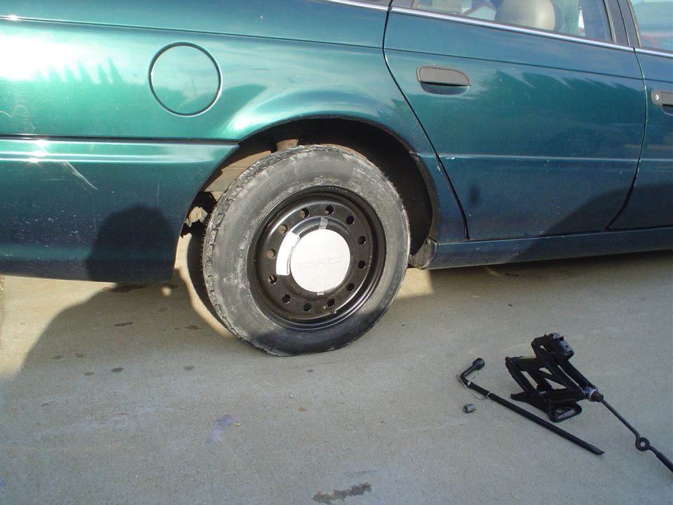 Green 1993 Ford Taurus sedan with a flat rear tire, jack, and lug wrench on concrete
