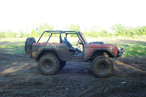 1974 Ford Bronco - A mud-covered red early Ford Bronco photographed in profile, stripped to an open body with a tube roll cage, full doors removed, and oversi…
