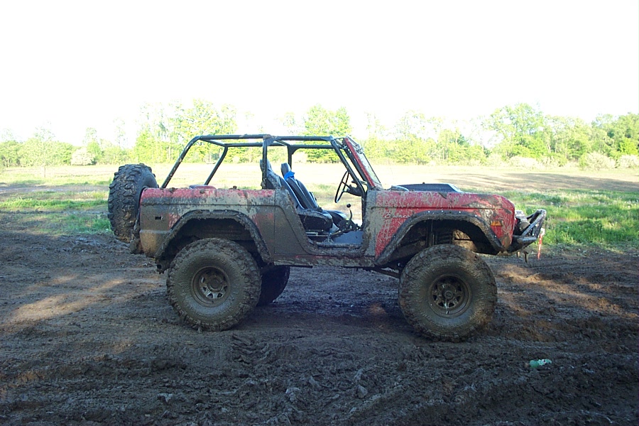 Muddy red lifted Ford Bronco with roll cage and big off-road tires, side view in a field.