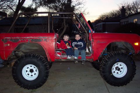 1974 Ford Bronco - Side view of a red early Ford Bronco stripped of top and doors, fitted with a full roll cage, lifted suspension, and large mud tires on whi…