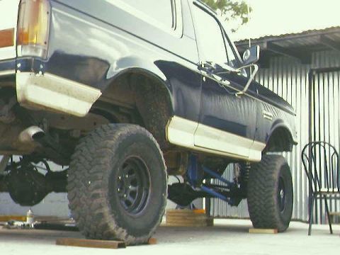 1990 Ford Bronco - The rear suspension of a lifted Ford Bronco is visible, showing custom blue components and large off-road tires.