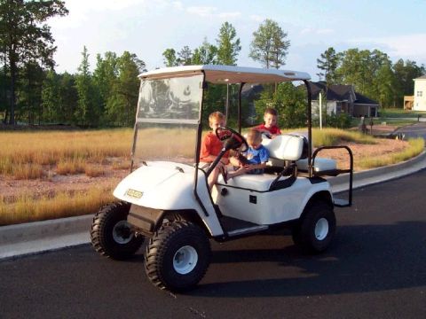 1979 Ford Bronco - A white golf cart carries three young children on a suburban street.