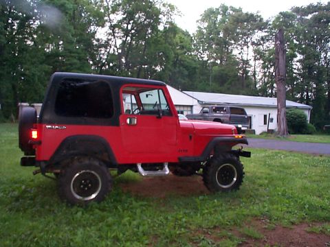 1989 Jeep YJ - A red Jeep Wrangler is parked on the grass in front of a house with a dark-colored truck in the driveway.