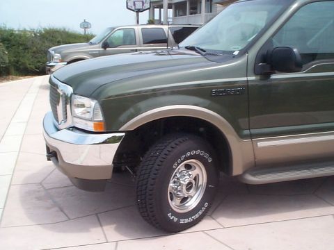 2001 Ford Excursion - Two Ford Excursions are parked on a paved area near a building and some greenery.