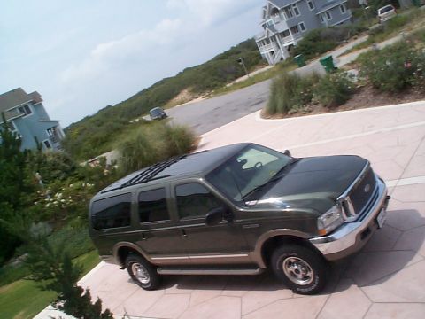 2001 Ford Excursion - A dark green 2001 Ford Excursion is parked on a paved driveway, with coastal houses and vegetation visible behind it.