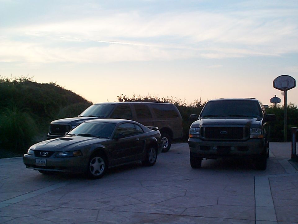 Dark Mustang coupe, Ford Excursion SUV, and Ford Super Duty pickup parked on driveway at dusk