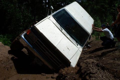 1988 Ford Bronco - A white SUV is stuck in a muddy ditch, with a person attempting to push it out.