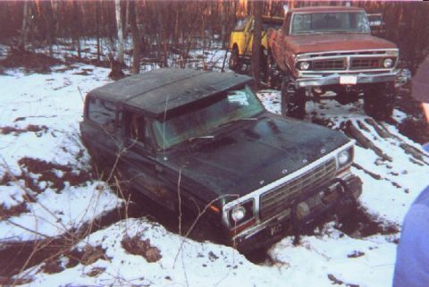 1976 Ford F-250 - A dark SUV is stuck in a snowy ditch, with a red Ford pickup truck visible in the background.