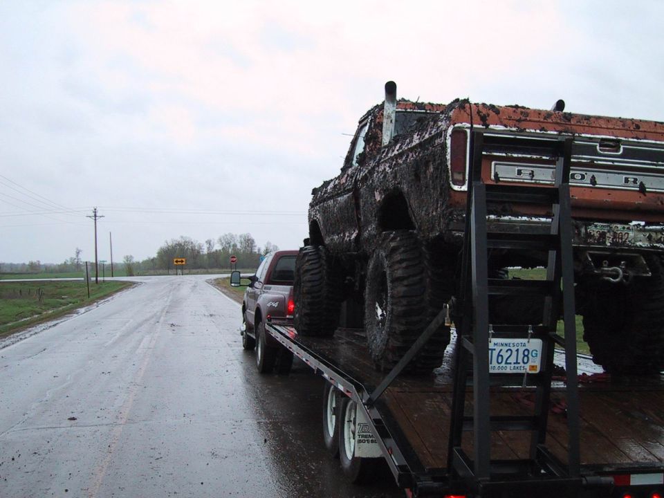 Red Ford pickup towing a muddy monster-style Ford truck on a flatbed trailer on a wet road.