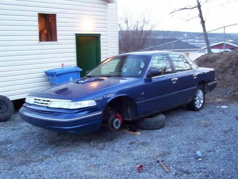 1995 Ford Crown Victoria - A blue 1995 Ford Crown Victoria is parked outside a building with its front wheels off.