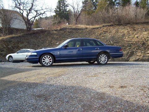 1995 Ford Crown Victoria - A blue 1995 Ford Crown Victoria is parked on a gravel lot next to a white car.