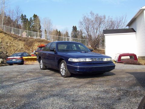 1995 Ford Crown Victoria - A blue sedan is parked in a gravel lot next to a white building, with another vehicle and equipment in the background.