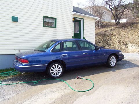 1995 Ford Crown Victoria - A blue 1995 Ford Crown Victoria sedan is parked next to a house with a green garden hose on the ground.