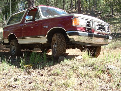 1990 Ford Bronco - A red and white two-tone late-1980s Ford Bronco XLT posed at an angle on a rutted dirt track in a pine forest, seen from the front three-qu…