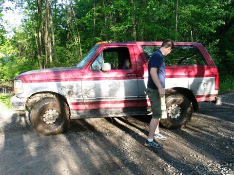 1995 Ford Bronco - A man stands beside a red and silver Ford Bronco on a dirt path surrounded by trees.