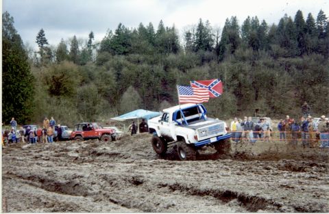 1990 Ford Bronco - Off-road trucks compete in a mud bog event with spectators watching.