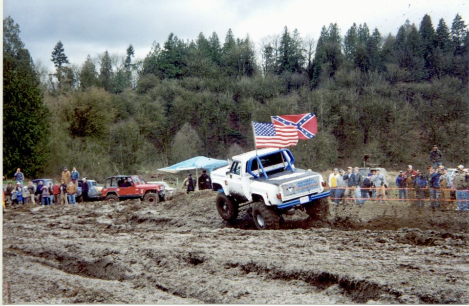 White off-road truck with blue roll cage driving through mud, American and Confederate flags flying