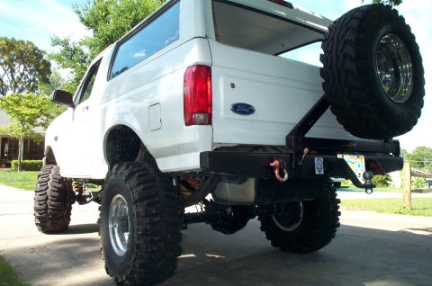 1995 Ford Bronco - Low rear three-quarter view of a heavily lifted white Ford Bronco on tall Super Swamper-style tires, with a black tube rear bumper, red D-r…
