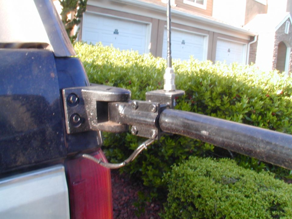 Close-up of a metal bracket with an antenna mount attached to a dark blue vehicle body. A black pipe extends horizontally
