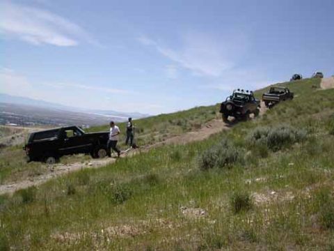1994 Ford Bronco - Multiple off-road vehicles navigate a dirt track on a grassy hillside with a distant city view.