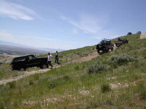 Several off-road vehicles driving up a grassy hill on a dirt track under a blue sky