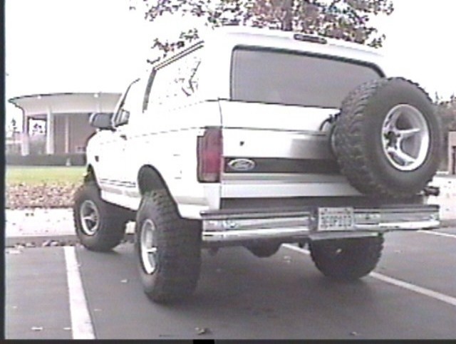Lifted white Ford Bronco with rear-mounted spare tire, parked in a parking lot, rear three-quarter view