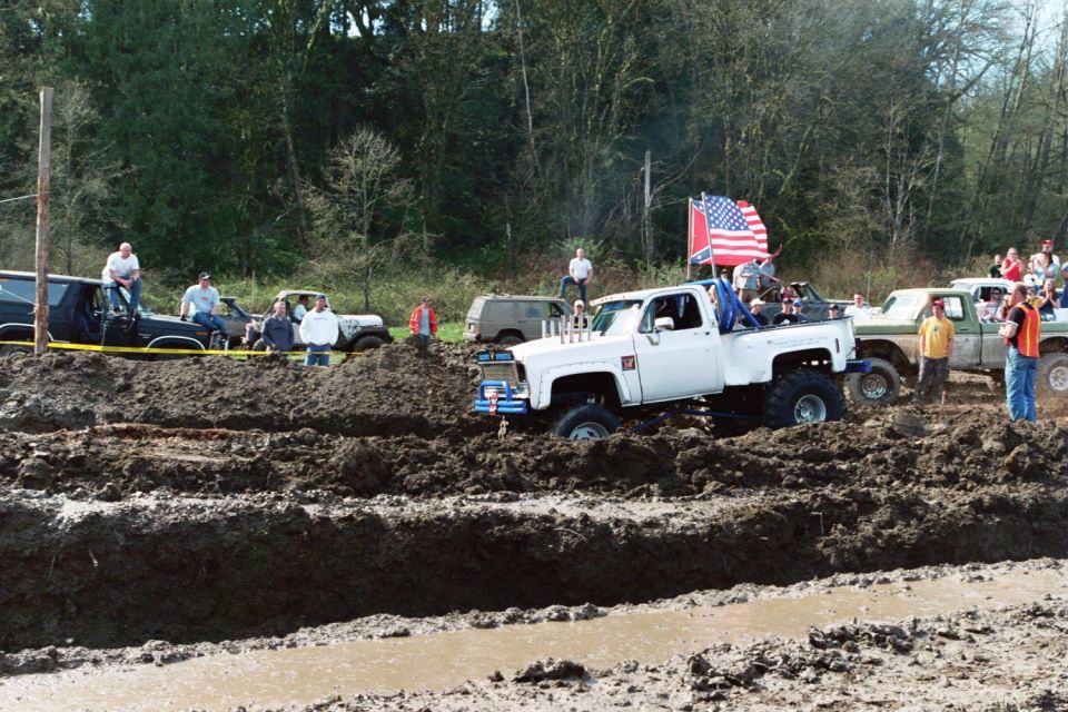 White pickup truck with large tires navigating a mud pit, American and Confederate flags flying