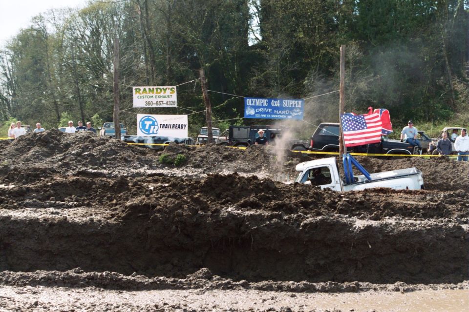 White pickup truck stuck in mud pit, American flag on pole behind it. Other vehicles and spectators visible