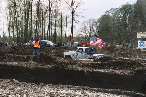 1990 Ford Bronco - A white pickup truck is stuck in a mud pit with spectators watching and flags flying.