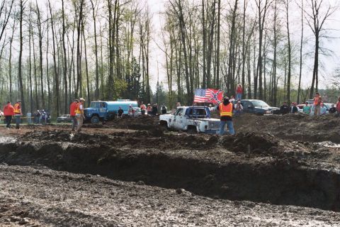 1990 Ford Bronco - A muddy scene with several vehicles and people participating in an off-road event, flags.