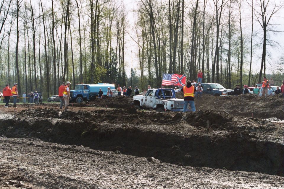 Mud bogging scene with white pickup truck stuck in mud, American and Confederate flags flying